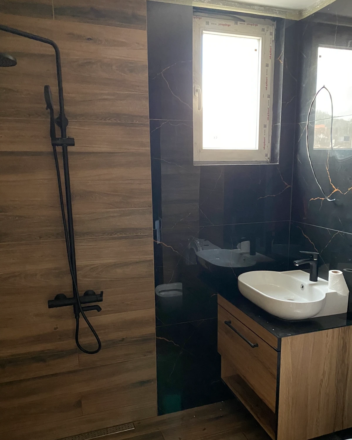 Bathroom in black marble and wood-look tiles: black shower set, basin on a wooden vanity unit, window